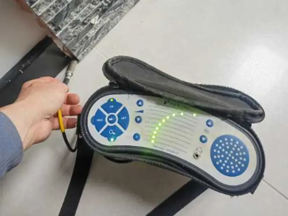 Tracer gas analyser held at floor level near tiled wall during leak detection